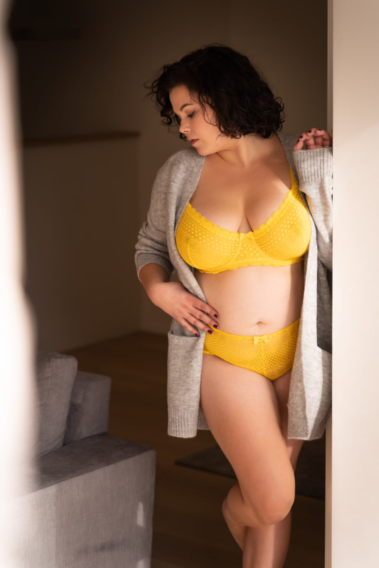Femme en lingerie jaune et gilet gris appuyée contre un encadrement de porte